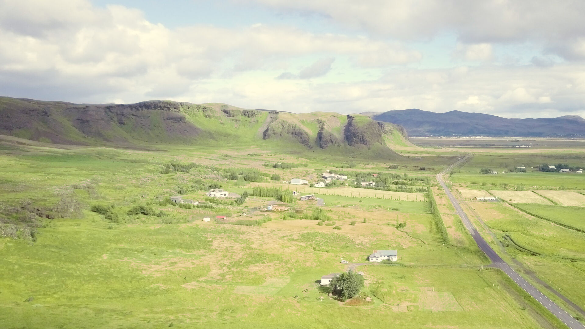 Aerial view of Riftún in Ölfus, Iceland: green meadows below lava cliffs, the farmhouse and road 38